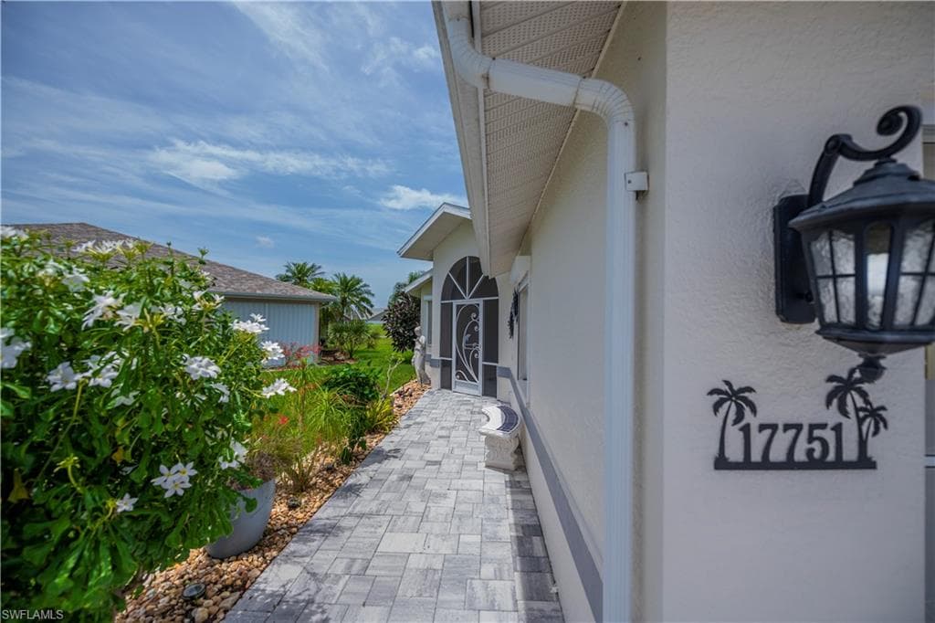 Exterior walkway featuring paver stone, lush landscaping, a decorative outdoor light fixture, and house numbering