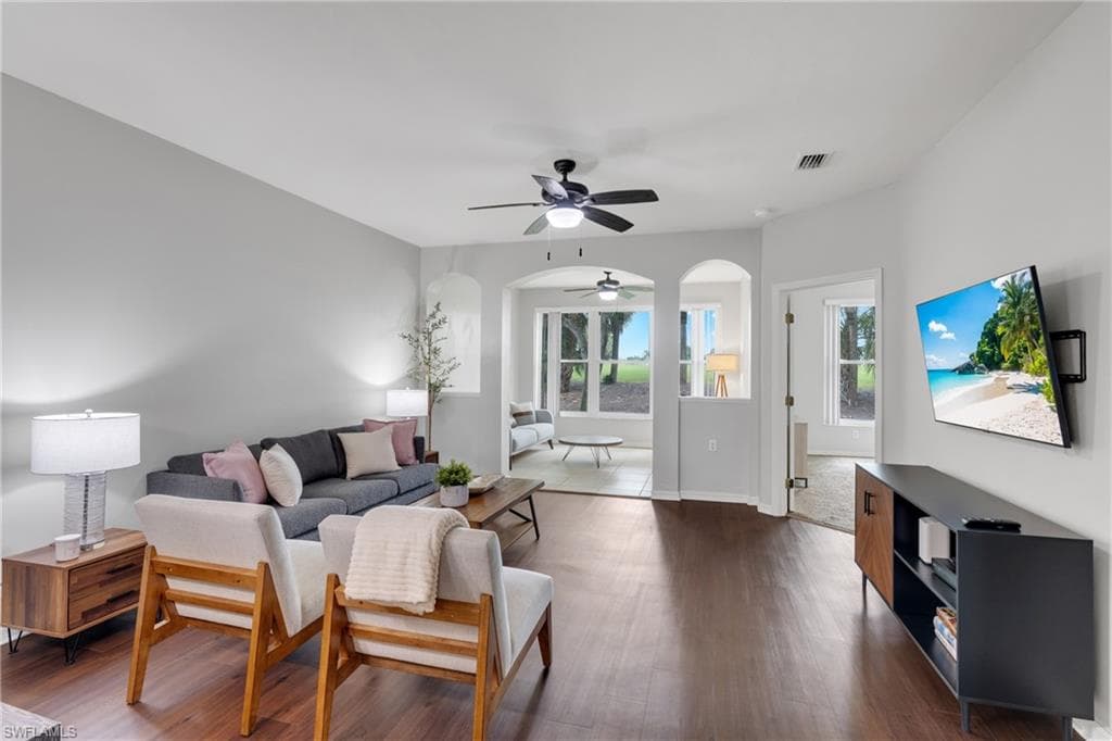 Spacious living area featuring rich wood-finish flooring, light gray walls, and an arched entry to an additional sunroom