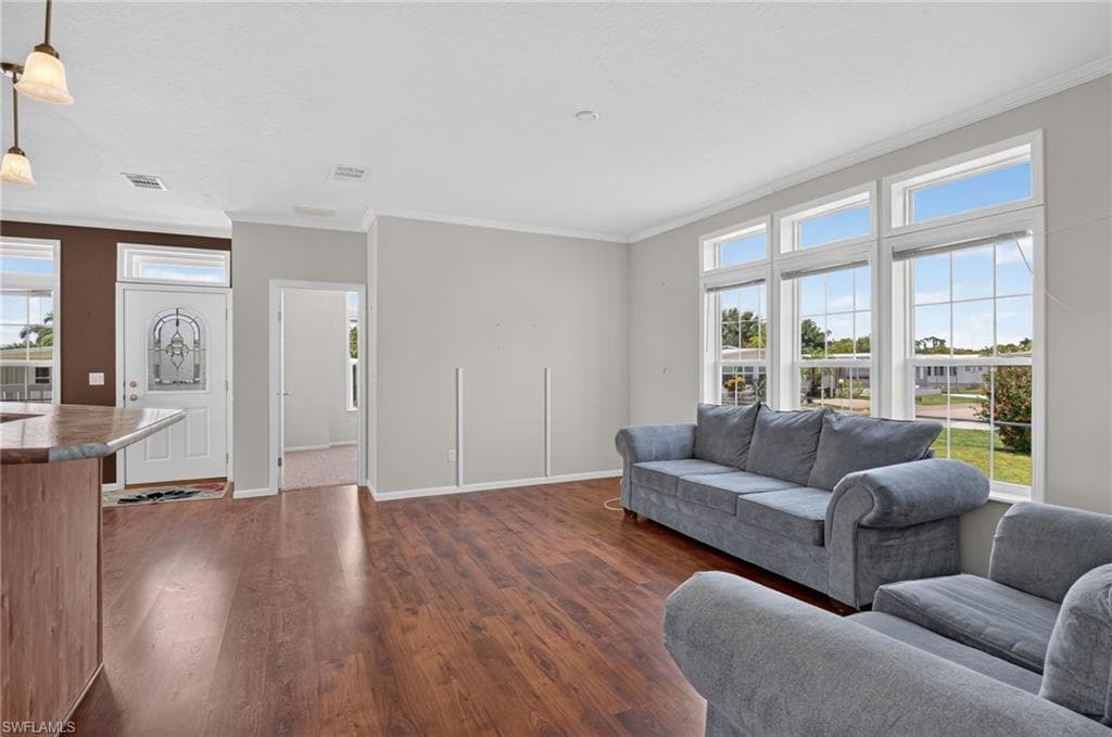 Bright living area featuring wood-finish flooring, a large window array with divided lites, crown molding, and a partial view of a kitchen island with a stone-look countertop
