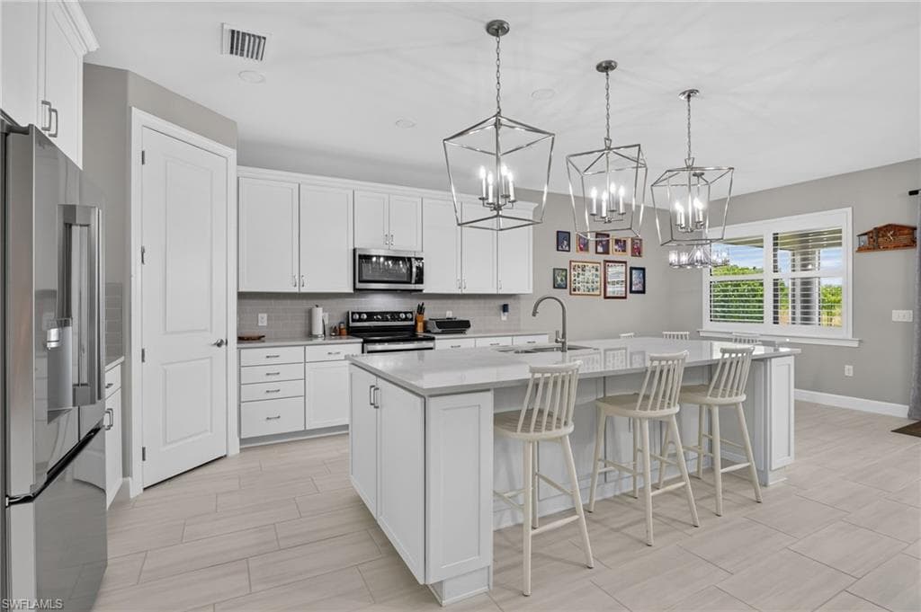 Spacious kitchen featuring white cabinetry, light countertops, stainless steel appliances, a center island with sink, and three geometric pendant lights