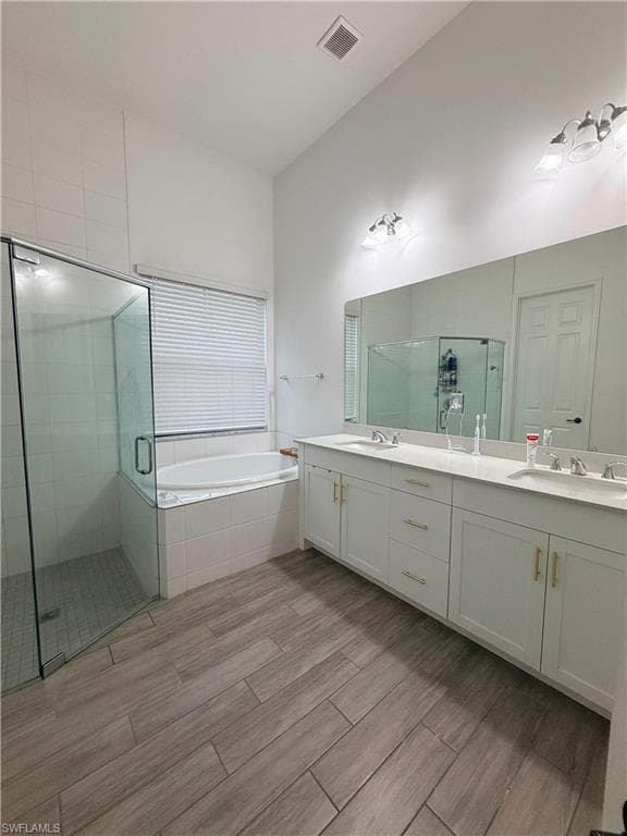 Bathroom featuring a dual vanity with white cabinetry and gold-tone hardware, a large frameless mirror, a glass-enclosed shower, a corner soaking tub, and wood-finish tile flooring