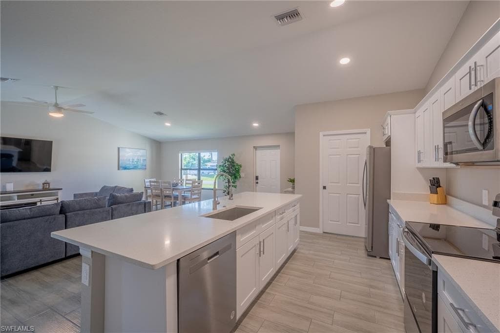 Open-concept kitchen featuring a large island with an undermount sink, light-toned countertops, white cabinetry with brushed nickel hardware, stainless steel appliances, and wood-finish tile flooring