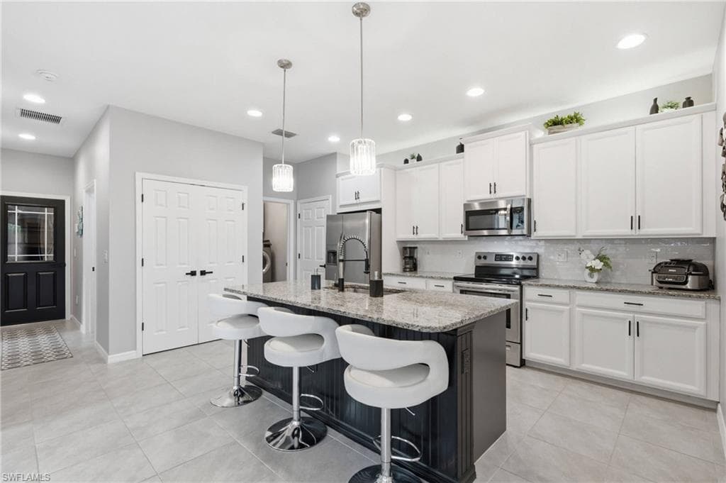 Modern kitchen featuring a granite-topped island with pendant lighting, stainless steel appliances, white cabinetry with black hardware, a tiled backsplash, and light-toned tile flooring