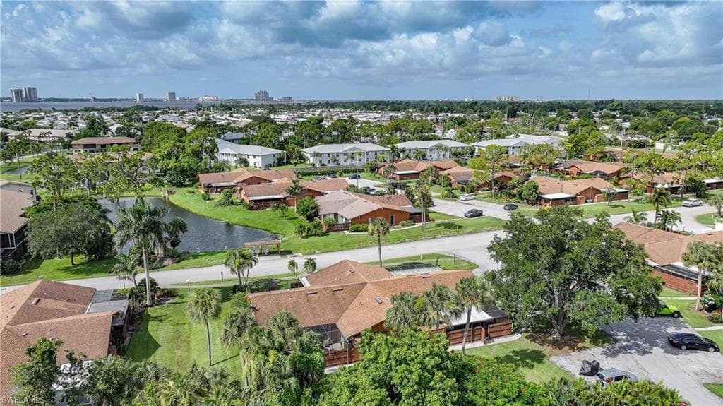 Residential community featuring a prominent central pond, mature palm trees, diverse architectural styles, and paved roadways, all set against a distant urban skyline