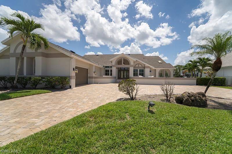Expansive paver driveway leading to a residence featuring a stucco exterior with decorative banding, arched windows, and mature palm trees