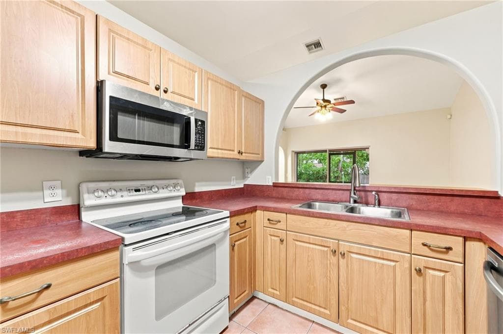 Kitchen featuring light wood cabinetry, a stainless steel over-the-range microwave, a white electric range, a double basin stainless steel sink, and red countertops