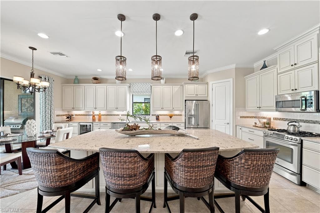 Spacious kitchen featuring an oversized island with a light-toned countertop, white cabinetry with crown molding, stainless steel appliances, and pendant lighting