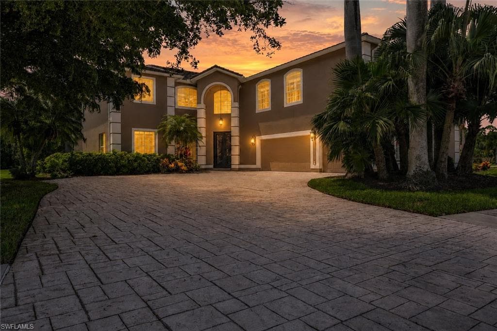 Stucco exterior with architectural details, arched entry, two-car garage, mature landscaping, and a paver driveway