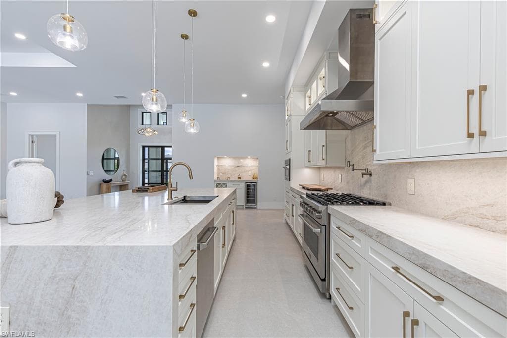 Spacious kitchen featuring a large waterfall island with integrated sink, white shaker-style cabinetry with gold-tone hardware, stainless steel appliances, a six-burner gas range with a pot filler, and integrated wine refrigeration
