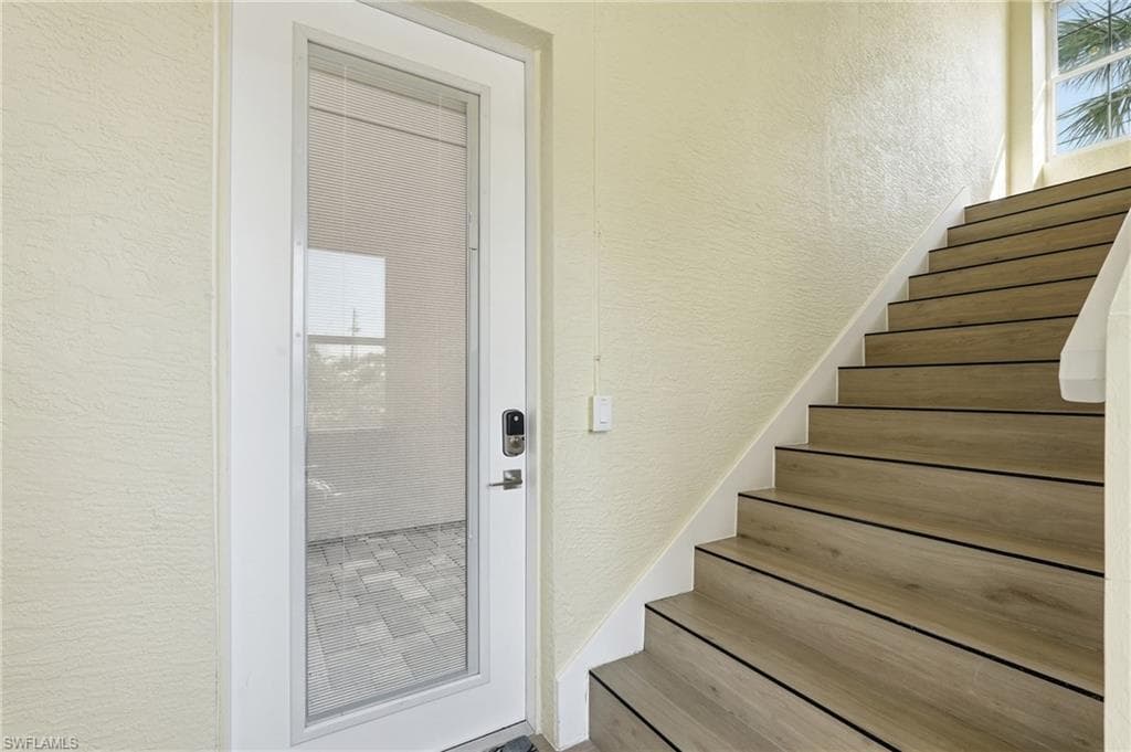 Full-lite glass door with a built-in smart lock, wood-finish staircase with white risers, textured wall finish, and upper-level window providing natural illumination - Virtually Edited Image