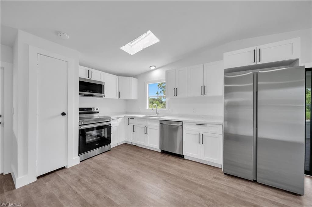 Modern kitchen featuring white cabinetry, stainless steel appliances, a white solid surface countertop, wood-finish flooring, and a ceiling-mounted skylight