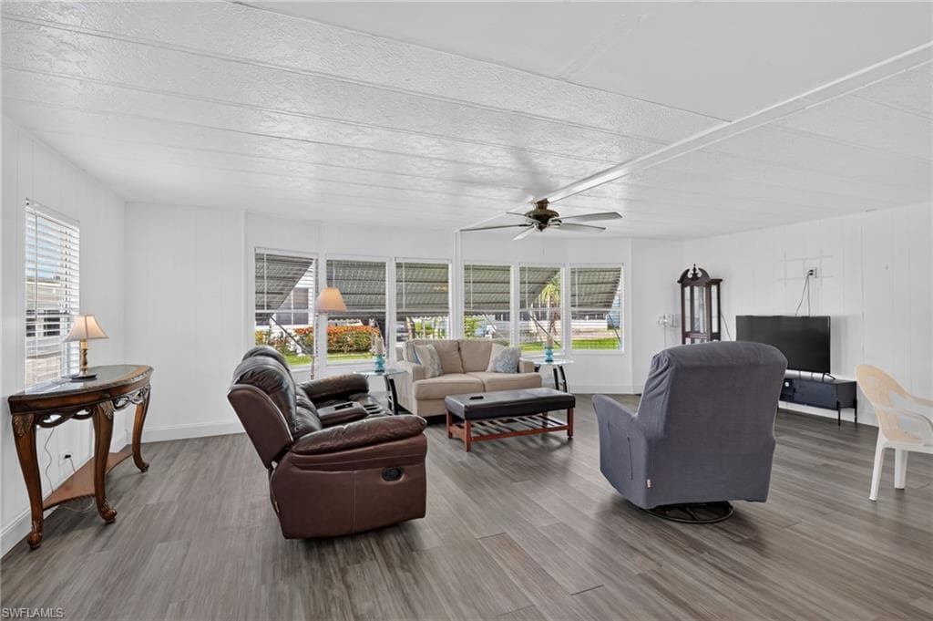 Spacious living area featuring wood-finish flooring, white paneling, multiple windows with blinds, and a ceiling fan with light fixture