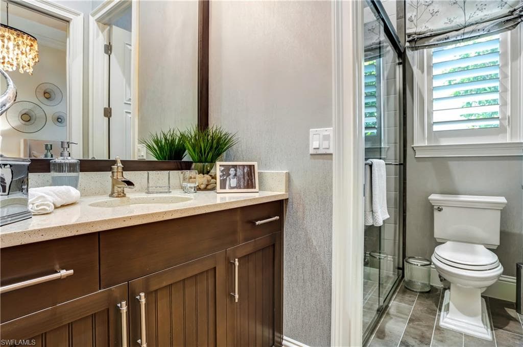 Bathroom featuring a dark wood-finish vanity with a light-toned countertop, a frameless glass shower enclosure, a window with plantation shutters, and large format floor tiling