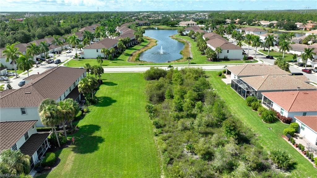 Expansive cleared lot bordered by lush green landscaping and tropical foliage, situated within a community featuring a central water body with a fountain