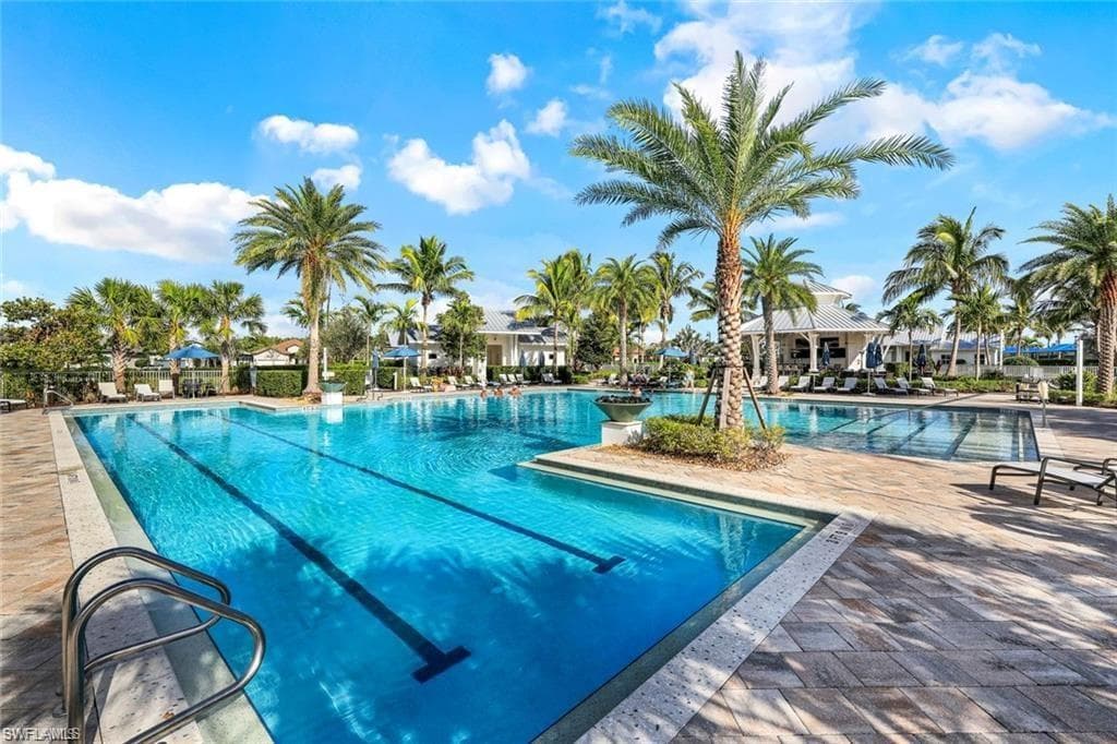 Resort-style pool area featuring a large lap pool with lane markers, a secondary pool, extensive paver decking, abundant palm trees, and a clubhouse structure