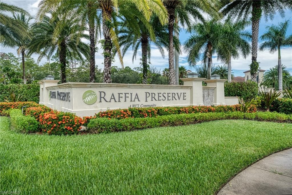 Raffia Preserve community entrance featuring a stone monument sign, lush green lawn, vibrant flowering shrubs, and tall palm trees
