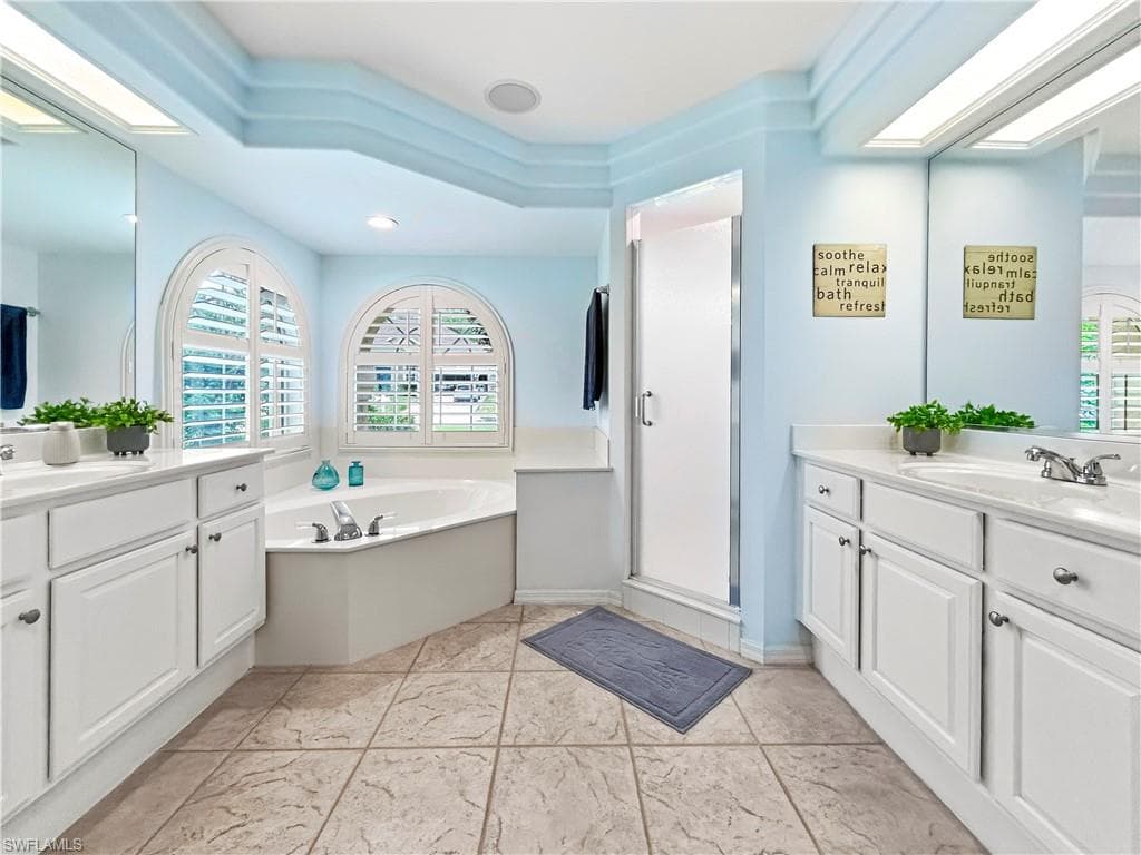 Dual vanities with white cabinetry, a corner soaking tub, two arched windows with plantation shutters, a glass-enclosed shower, and textured tile flooring