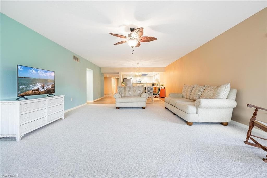 Spacious living area featuring light-colored carpeting, a ceiling fan with integrated lighting, and a partial opening to the kitchen