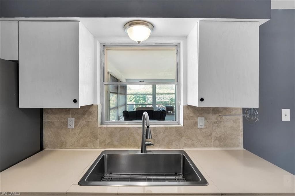 White upper cabinetry, stainless steel basin sink with a pull-down faucet, light-toned countertop, and a tiled backsplash