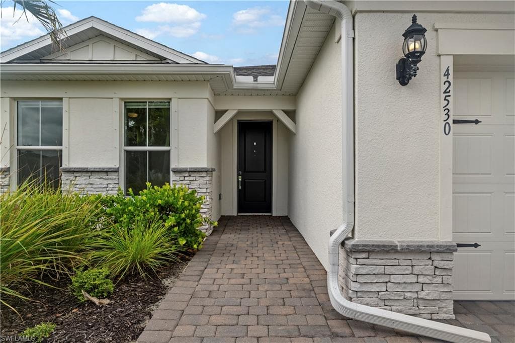 Paver walkway leading to an entryway with a black door, exterior wall light, and a two-car garage