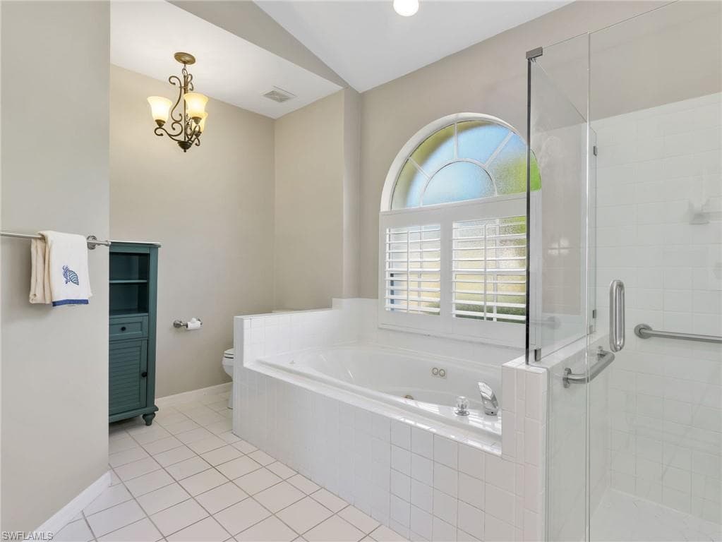 Soaking tub with white tile surround, arched window featuring plantation shutters, glass-enclosed shower, and tile flooring