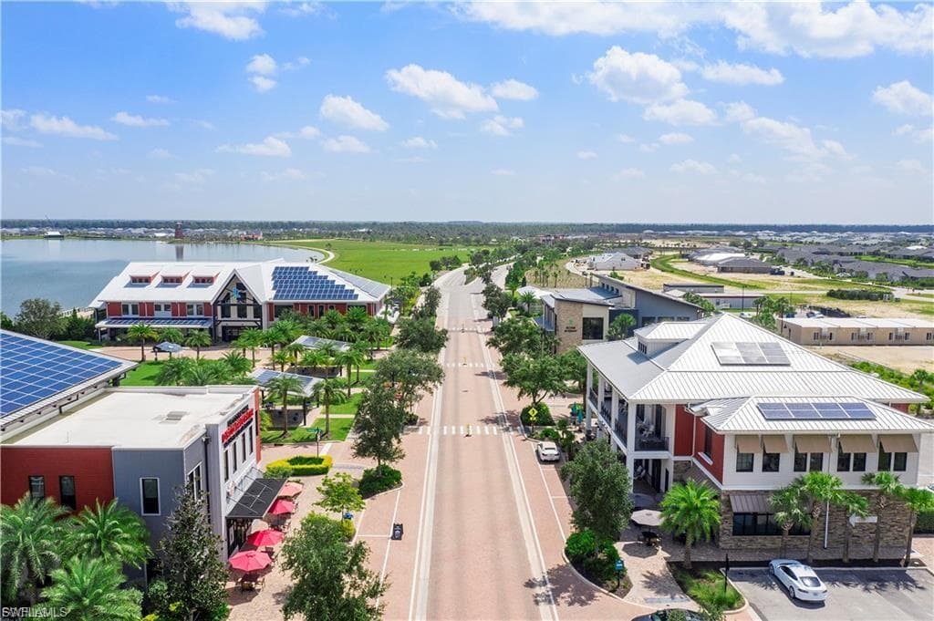 Aerial view showcasing a modern residential community featuring contemporary architecture with mixed red and white exterior finishes, solar panel installations, manicured landscaping with mature trees lining a central boulevard, and expansive green spaces overlooking agricultural countryside and waterfront in the distance