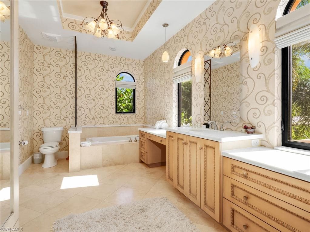 Vaulted ceiling with an ornate chandelier, dual arched windows, built-in soaking tub, extensive vanity with dual sinks, and tile flooring