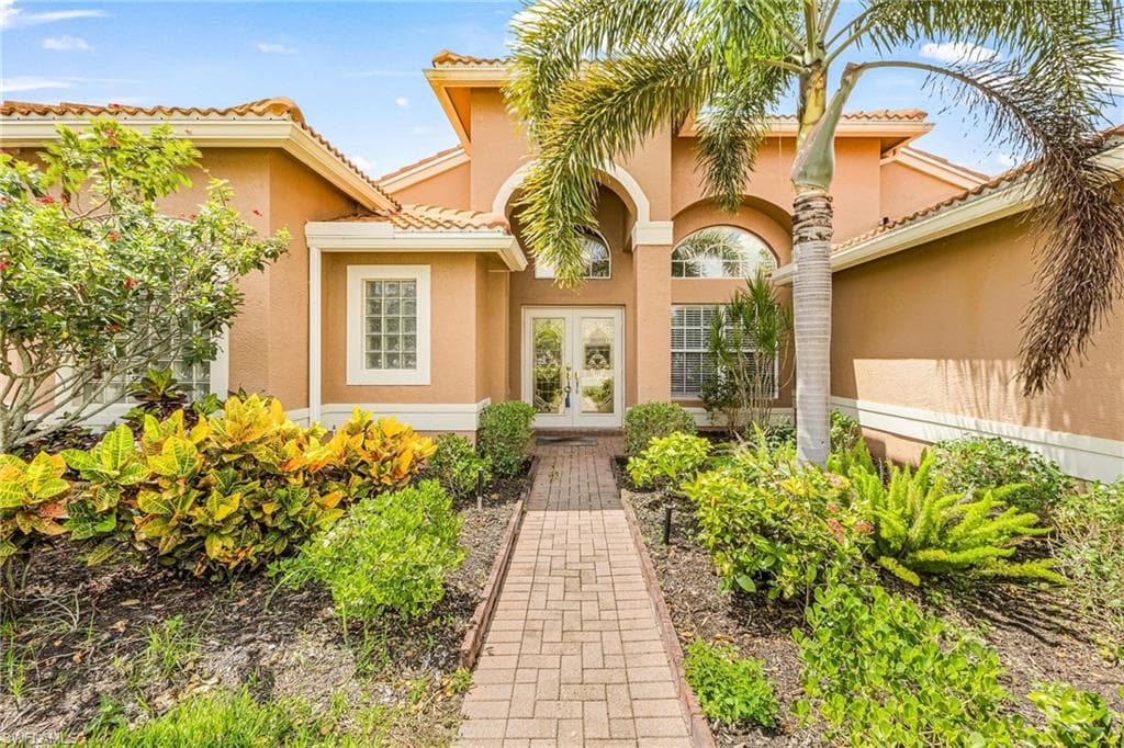 Stucco exterior with a tile roof, dual glass-paneled entry doors, an arched transom window, a block-patterned pathway, and diverse landscaping