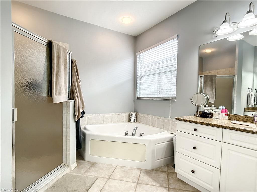 Corner soaking tub, separate shower with frosted glass enclosure, dual vanity with granite-finish countertop, white cabinetry, and tiled flooring