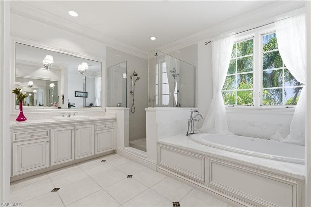 White tiled flooring with dark accent squares, double vanity with white cabinetry and integrated sinks, large mirrored wall, glass-enclosed shower, and a built-in tub with a window