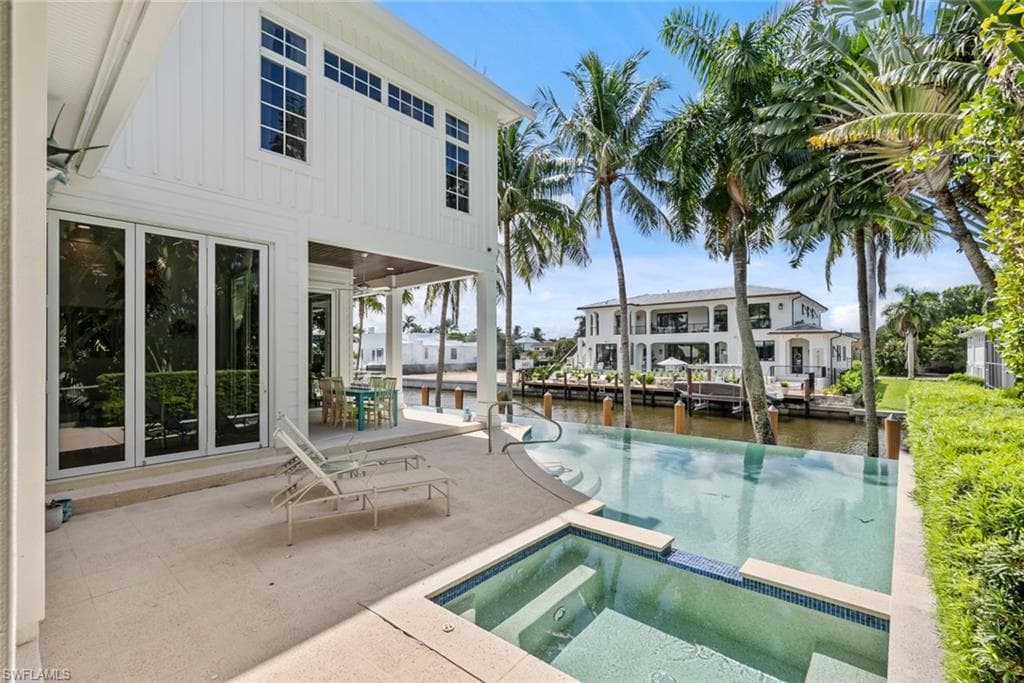 White vertical siding exterior with expansive multi-panel glass doors, covered patio, infinity-edge pool, and integrated spa with mosaic tile accents