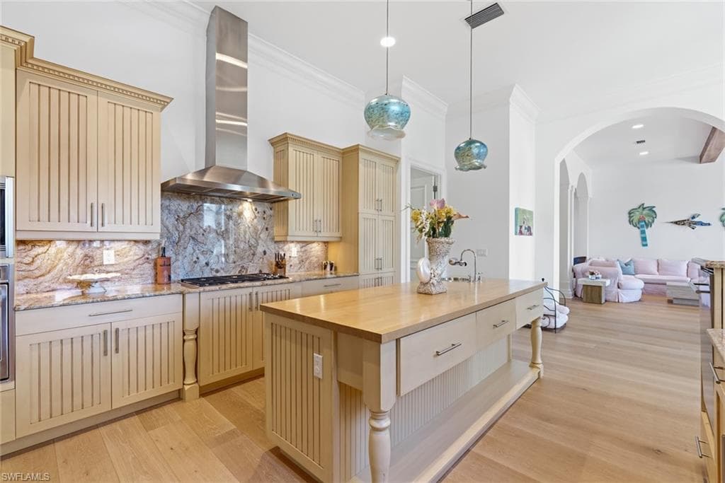 Kitchen featuring light wood-finish cabinetry, stone slab backsplash, stainless steel range hood, and a light wood-finish island with integrated storage