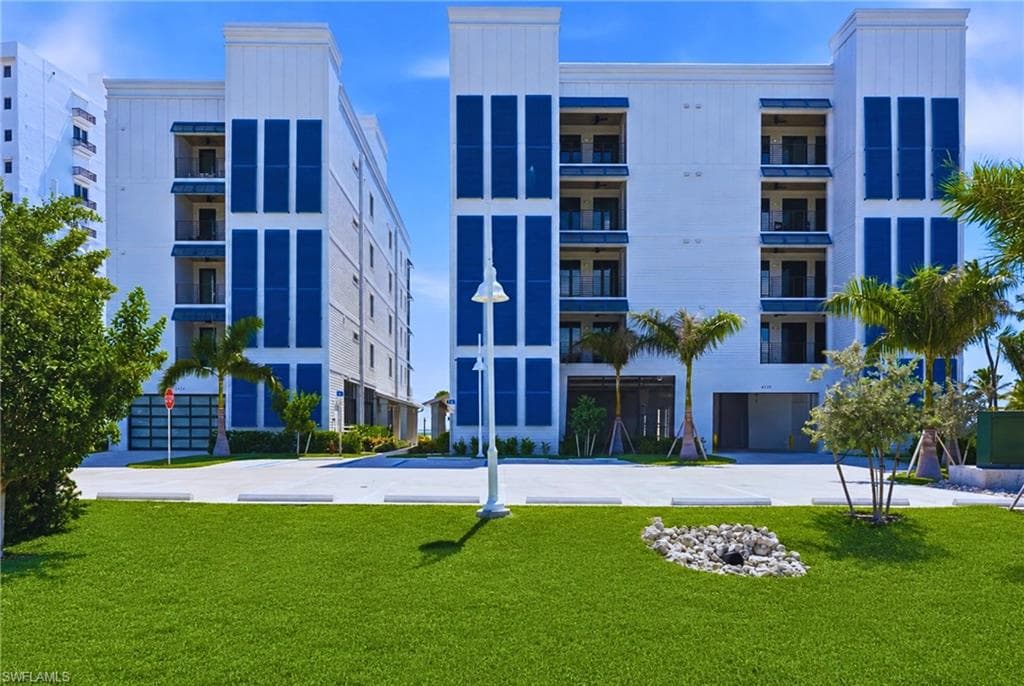 Multi-story building featuring white siding and blue accents, private balconies with black railings, ground-level parking garage, and a manicured lawn - Virtually Edited Image