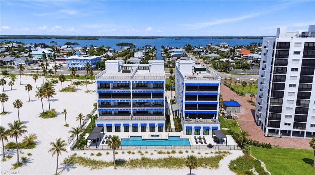 Coastal condominium architecture with multiple private balconies, a resort-style swimming pool, surrounding sandy beach, and mature palm treesExpansive land parcel with direct beach access, surrounded by established buildings and roadways - Virtually Edited Image