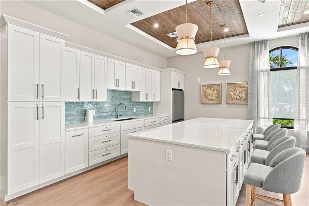 White shaker cabinetry, light wood-finish flooring, wood plank ceiling accent, large kitchen island with integrated seating, and a tiled backsplash