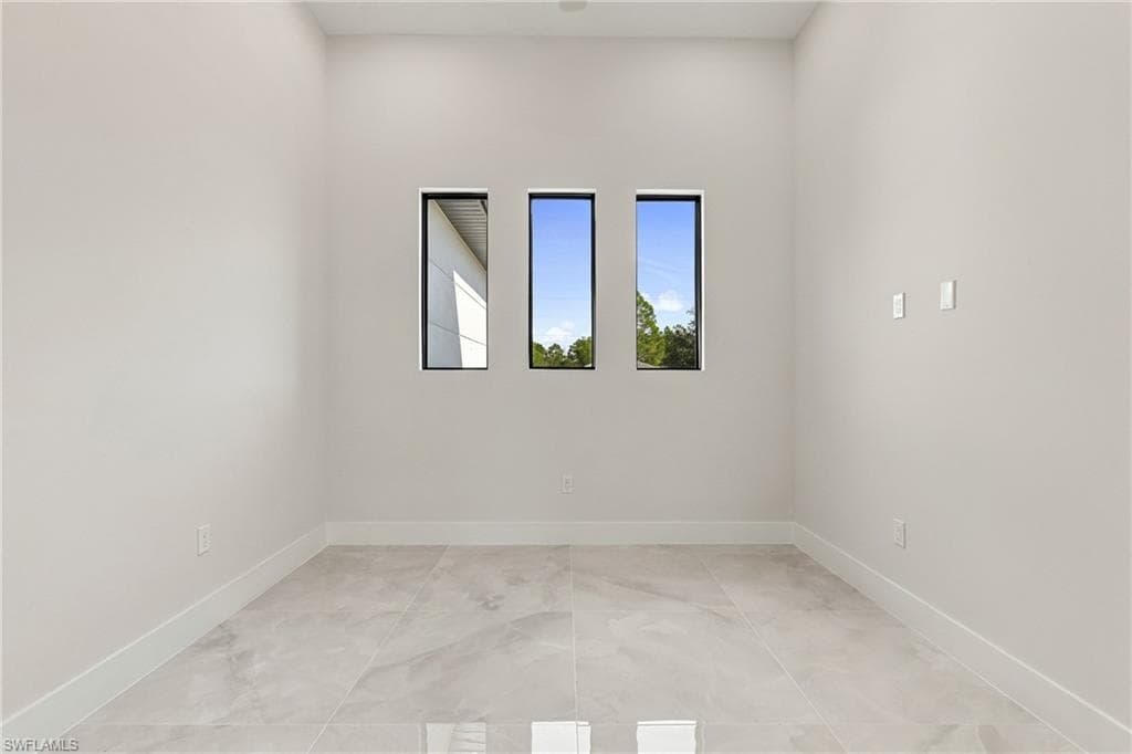 Recessed lighting illuminates a room featuring three vertical windows with dark frames, light-toned walls, and large format floor tiling