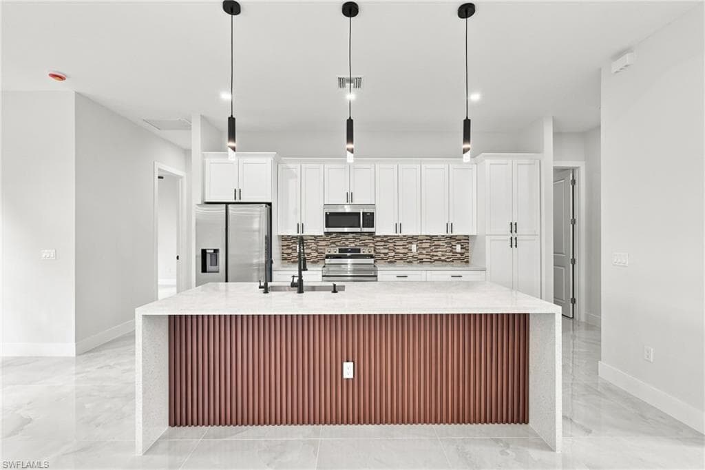 Kitchen island with waterfall countertop, white cabinetry, integrated stainless steel appliances, mosaic tile backsplash, and three pendant light fixtures