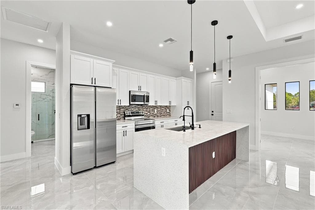 White shaker cabinetry with black hardware, stainless steel appliances, patterned tile backsplash, quartz countertop island with wood-finish paneling, and pendant lighting