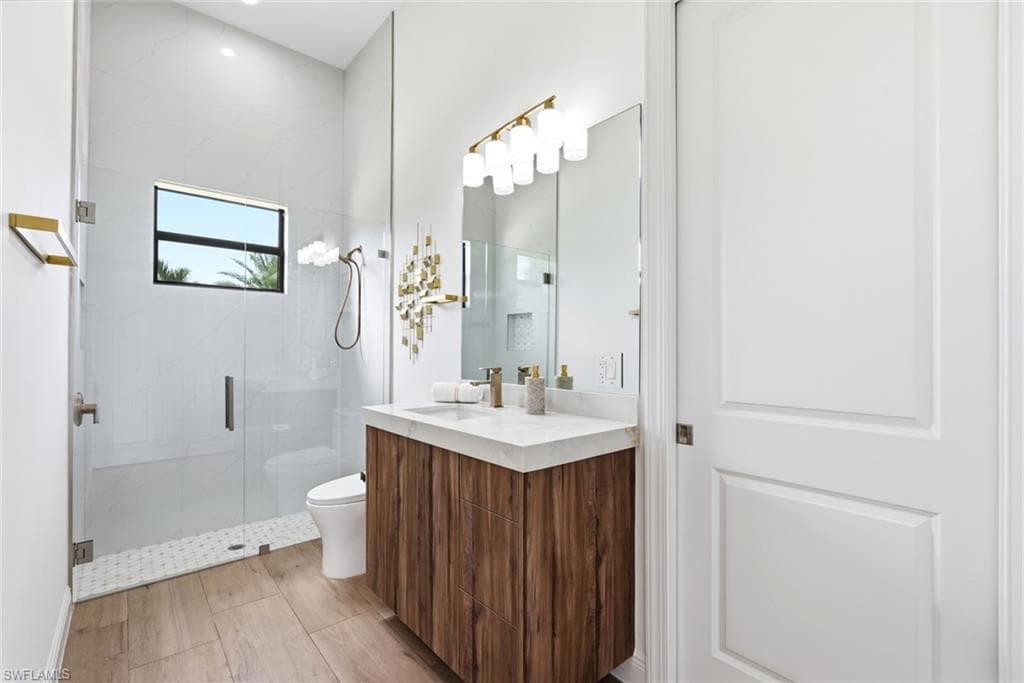 Frameless glass shower enclosure, wood-finish flooring, floating vanity with a light-toned countertop, modern vanity lighting, and a window