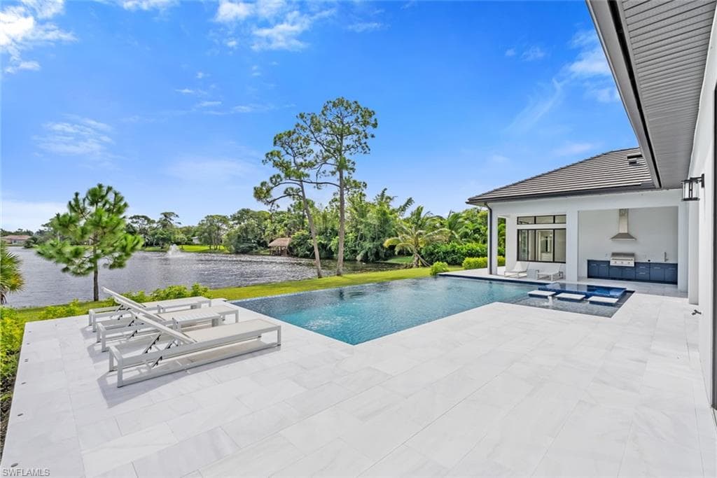 Infinity-edge pool with an integrated spa, expansive white tile patio, and outdoor kitchen featuring built-in cabinetry and a stainless steel range hood