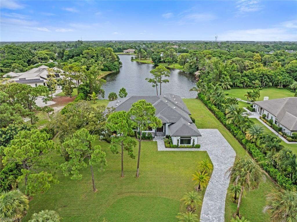 Waterfront property featuring a paver driveway, lush green lawn, mature trees, and a tile roof
