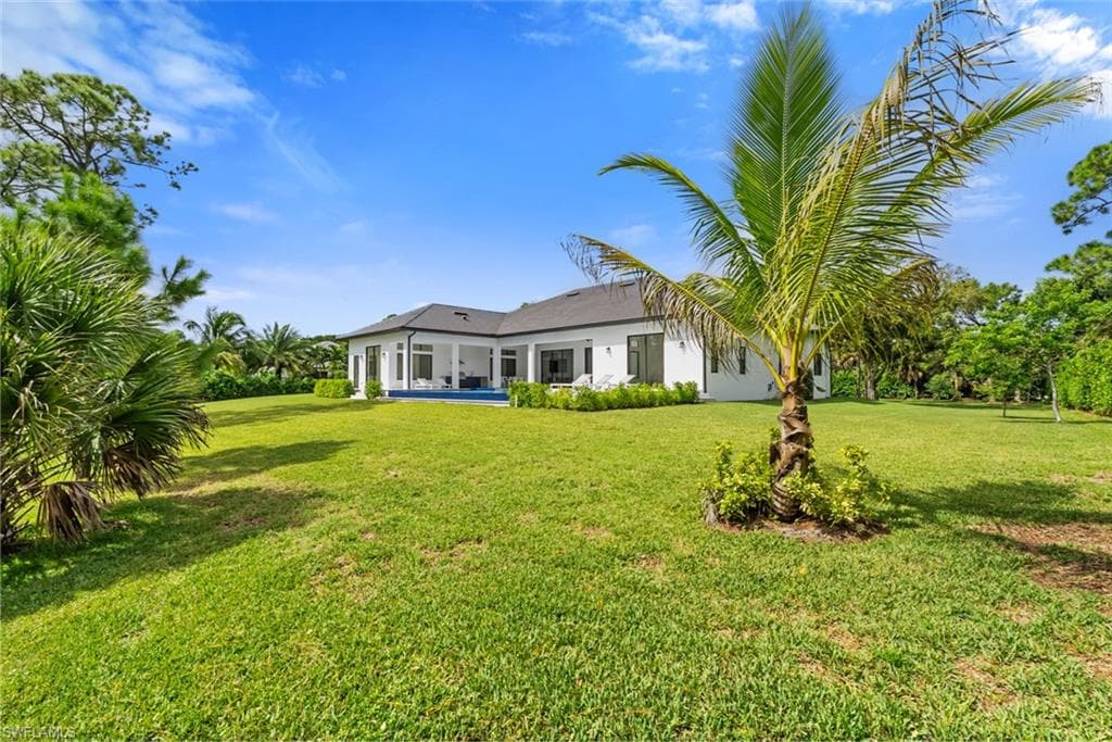 Expansive green lawn with mature trees and a palm tree, featuring a white exterior residence with a dark roof and covered patio