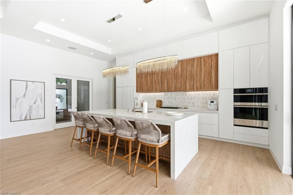 Expansive kitchen island with a waterfall edge, integrated double wall ovens, wood-finish upper cabinetry, and recessed ceiling lighting